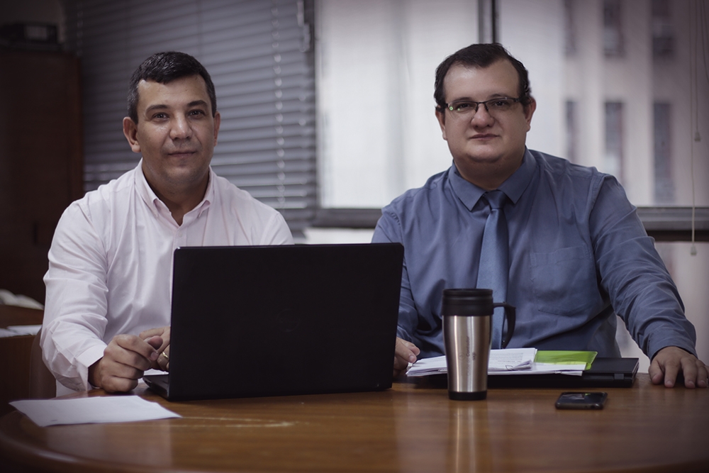 Os sócios Renato e Rodrigo, em frente a uma mesa com computador e outros objetos.